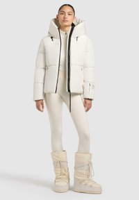 Giacca puffer bianca con cappuccio alto, zip nera e tasche laterali; abbinata a leggings color crema e stivali da neve bianchi con lacci.