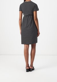 Femme portant une robe noire et blanche à rayures horizontales, longueur genou, manches courtes, debout de dos, portant des chaussures à talons blocs noires.