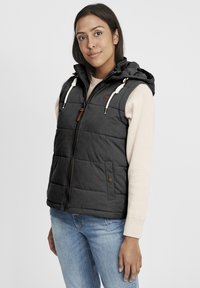 Svart vadderad väst med huva, med dragkedjestängning, sidofickor och vita dragsnören. Bärs över en ljus tröja och jeans.