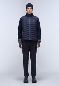 Marinblå puffväst med quiltat mönster och blå dragkedja, kombinerad med mörka byxor och fingerlösa grå vantar, samt en stickad marinblå mössa.