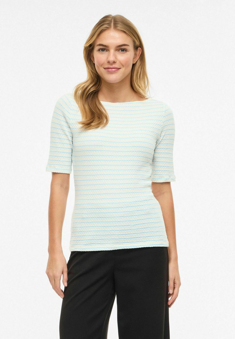 Hosszú szőke hajú nő, világoskék és fehér csíkos rövid ujjú felsőt és fekete nadrágot visel, egyszerű fehér háttér előtt áll.