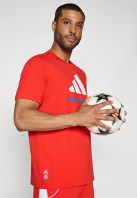 Rød t-skjorte med korte ermer og hvit Adidas-logo samt blå tekst "BAYERN MÜNCHEN". modellen holder en svart og hvit fotball. Minimalistisk design.