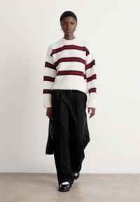 Hvit strikket genser med røde og svarte striper, ribbestrikket kant; brukt med løse svarte bukser og svarte sko, minimalistisk design, modell som går.
