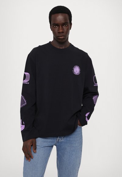 Schwarzes Langarm-T-Shirt aus Baumwolle mit lila Grafikdruck auf Ärmeln und Brust. Hellblaue Jeans ergänzen das Outfit.