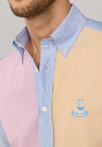 Man draagt een kleurgeblokte blouse met een blauwe kraag, roze en beige panelen, en een blauw geborduurd logo aan de beige zijde.