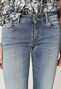 Jeans de denim azul claro con un lavado desgastado, diseño de cinco bolsillos y cierre frontal con botón. Detalles de costura sutil cerca de los bolsillos.