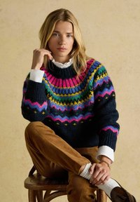 Pletený sveter s námorníckym telom, živý okrúhly yoke so vzormi v ružovej, zelenej a žltej farbe, skombinovaný s hnedými kordurovými nohavicami.