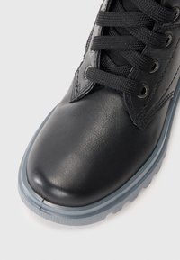 Zapato de cuero negro con puntera redondeada, superficie texturizada, cordones negros y suela de goma gris claro. Presenta ojetes plateados para los cordones.