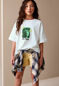 White t-shirt with green graphic of a face and five stars, paired with yellow shorts and a blue plaid shirt tied at the waist.