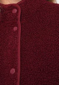 Suure plaan inimesele, kes kannab tumepunast fliiskatet, millel on pressnupud ja kare, puhvis kangas.