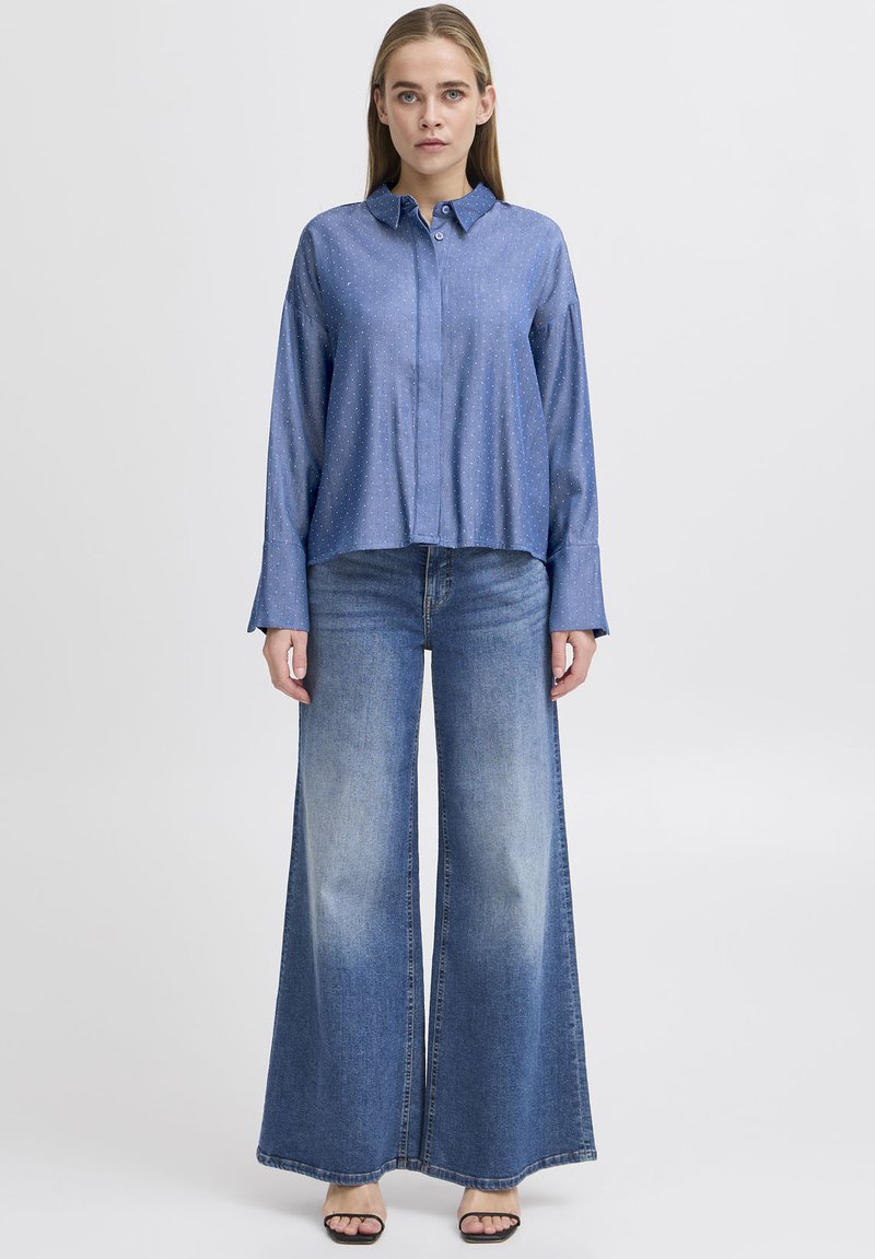 Lyseblå skjorte med knaplukning og et subtilt prikkemønster, parret med vidde denimjeans i en mørkere blå farve, begge lavet af glat stof.