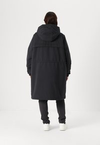 Long manteau noir rembourré avec une capuche, présentant un design matelassé et des poignets élastiques. Porté sur un pantalon gris et des baskets blanches.