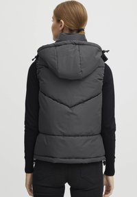 Gilet matelassé en charbon avec capuche amovible, présentant des motifs de diamants en losange et des cordons de serrage sur les côtés. Haut à manches longues noir en dessous.