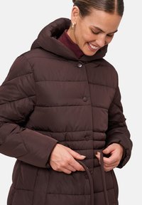 Bruine pufferjack met een aansluitende capuchon, drukknoopsluiting en gestikt patroon. Voorzien van een koord in de taille voor een verstelbare pasvorm.