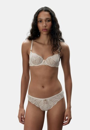 Weibliches Model mit langen lockigen Haaren, das passende cremefarbene Spitzen-BH und Slips trägt und vor einem schlichten hellen Hintergrund steht.