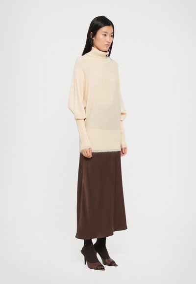 Beige neulepaita, jossa on leveät hihat, kerroksittain ruskean maksi-hameen päällä. Sujuva pinta, polvipituinen, yhdistetty tummiin korkokenkiin.