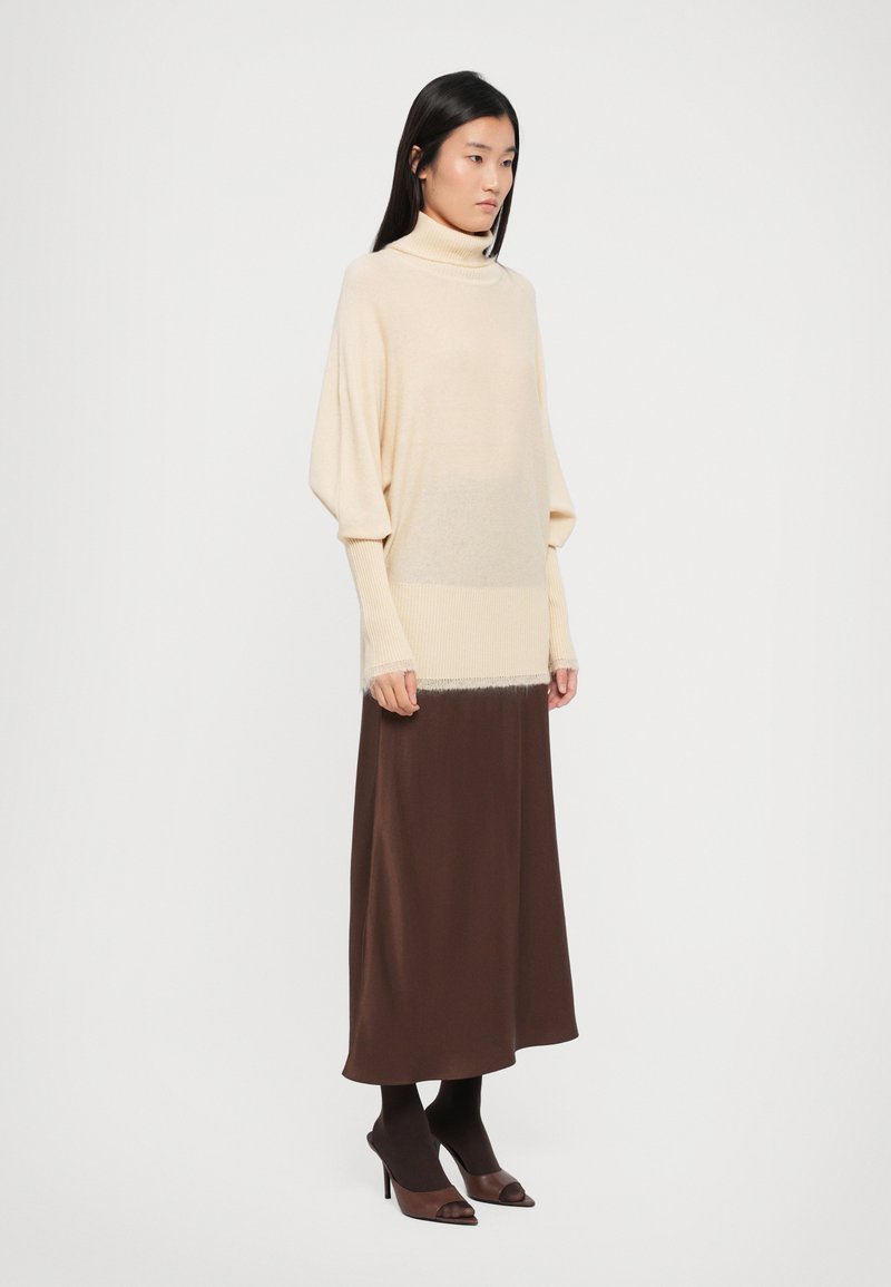 Un pull à col roulé beige en tricot avec des manches larges, superposé à une jupe maxi marron. Texture lisse, longueur genoux, associé à des chaussures à talons sombres.
