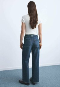 Brede denim jeans in donkerblauwe wassing met een hoge taille, voorzien van achterzakken en rafelige zomen. Gecombineerd met een aansluitende witte top.