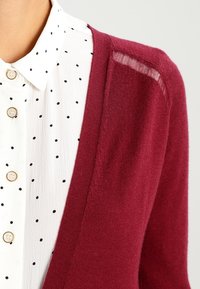 Bordový cardigan s měkkou texturou a otevřeným předním dílem, vrstvený přes bílou blůzu s černými puntíky a zlatými knoflíky.