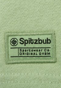 Hellgrüner Stoff mit einem eingenähten Etikett, auf dem in schwarzer Schrift "Spitzbub Sportswear Co. ORIGINAL OBM" steht, vor einem strukturierten Stoffhintergrund.