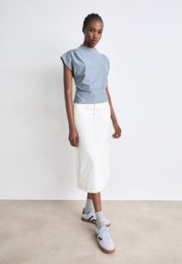 Femme debout contre un mur blanc, portant un haut à manches courtes bleu clair, une jupe midi blanche, des chaussettes grises et des baskets blanches avec des accents foncés.