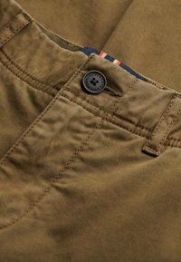 Pantalon en coton marron avec un bouton noir, des accents cousus et une ceinture bleue avec des rayures rouges. Détails de tissu texturé visibles.