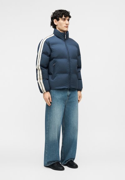 Mees, kes kannab tumel sinist pufferjopet valgete triipudega varrukatel, laia lõikega siniseid teksapükse ja musti jalanõusid, seisab ühevärvilise tausta ees.