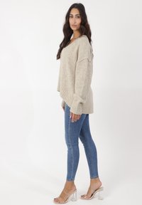 Suéter de punto oversized beige con puños acanalados, combinado con jeans skinny azules y sandalias de tacón bloque transparente, estilizado desde un lado.