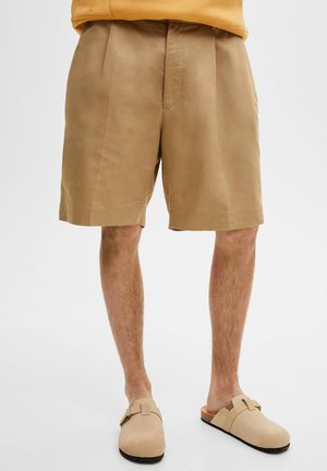 Shorts taillés beiges en tissu léger, dotés d'un devant plat, de poches latérales et d'une coupe décontractée, associés à des chaussures slip-on couleur tan.