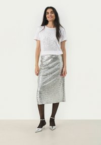 Camicetta bianca a maniche corte con decorazioni in borchie e una gonna luccicante di paillettes argentate. La modella indossa collant neri e décolleté argentati a punta.
