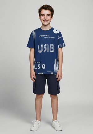 Lachende jongen die een blauw T-shirt met print, marineblauwe cargoshorts en witte sneakers draagt, staande tegen een effen achtergrond.