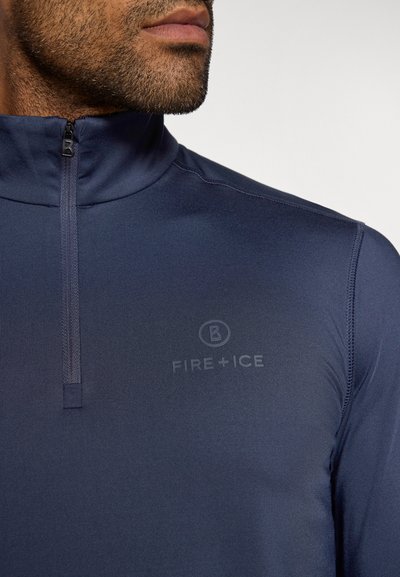 Close-up de um homem a usar um top azul-marinho com fecho de 1/4 e o logótipo "FIRE + ICE" no peito, com stubble de barba visível.