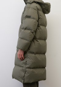 Side view of a person wearing a long, olive green, padded winter coat with a hood and snap buttons on the side hem.