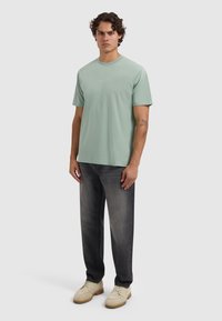 Man staat er in een lichtgroen T-shirt met korte mouwen, vervaagde zwarte jeans, beige schoenen en zichtbare tatoeages op zijn rechteronderarm.