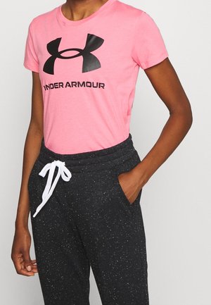 Osoba nosząca różowy t-shirt Under Armour i czarne spodnie dresowe z białym sznurkiem, z jedną ręką w kieszeni na jednolitym tle.