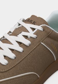 Καφέ σουέντ αθλητικό παπούτσι με λευκά κορδόνια, διάτρητο πλαϊνό πάνελ και το λογότυπο "ARKK" στη γλώσσα και στην εξωτερική πλευρά κοντά στη φτέρνα.