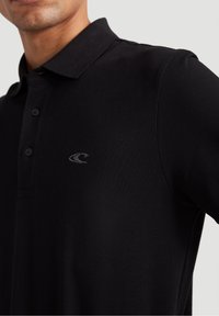 Muž v černém polokošili se třemi knoflíky a malým zakřiveným logem vyšitým na levé části hrudníku.