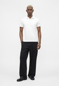 Homme aux cheveux blonds courts portant un polo blanc, un pantalon noir à jambes larges et des chaussures en cuir noires, debout devant un fond uni.