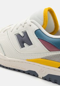 Baskets blanches New Balance avec des accents bleus, bordeaux et jaunes, semelle grise et col rembourré avec doublure jaune, montrant le logo et les lacets.