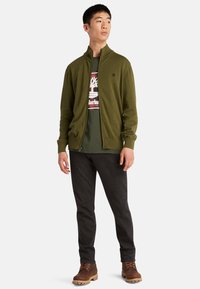 Grön zip-up jacka, mörka byxor, bruna stövlar. T-shirten har en grafisk design med en trälogotyp. Avslappnad, åtsittande silhuett med ribbade ärmmuddar.