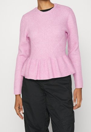 Personne portant un pull en maille texturée rose avec un basque à la taille et des manches longues, associé à un pantalon noir ample.