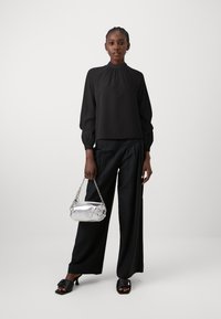 Blusa negra con cuello alto y mangas abullonadas, combinada con pantalones anchos negros y un bolso plateado con una correa de cadena. Sandalias negras de tacón cuadrado.