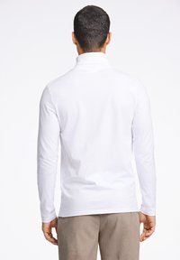 Baltas ilgomis rankovėmis polo marškinėlis su aukštu apykakle, lygiu paviršiumi ir įprasto silueto, derinamas su smėlio spalvos velveto kelnėmis.