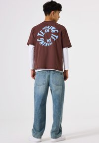 Camiseta marrón de manga corta con texto impreso en círculo de color azul en la parte de atrás, combinada con jeans baggy de color azul claro y zapatillas blancas.