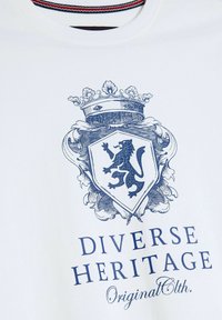 Biała bawełniana koszulka z nadrukiem w kształcie niebieskiego herbu z lwem, akcentami kwiatowymi oraz napisem "Diverse Heritage Original Cloth."