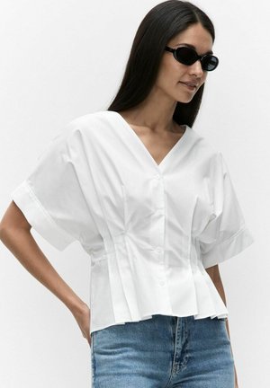 Vrouw met lang donker haar draagt zwarte ovalen zonnebril, een losse witte blouse met korte mouwen en knopen en blauwe jeans, staand tegen een witte achtergrond.