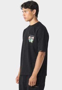 Młody mężczyzna w czarnej koszulce "Pas de Monaco" z kwiatowym i paskowanym wzorem tarczy, stojący w profilu na jednolitym tle.