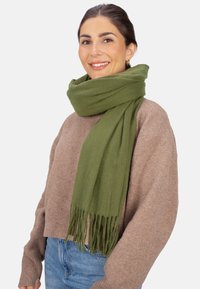 Écharpe vert olive en tissu doux, ornée de franges aux extrémités, portée sur un pull beige à coupe décontractée avec des poignets côtelés.