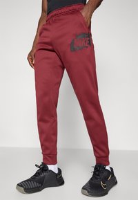 Burgundowe dresy z elastycznym pasem, z czarnym logo Nike na udzie, w połączeniu z czarnymi butami sportowymi i białymi skarpetkami.