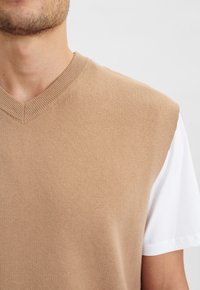 Beige ärmelloser Pullover mit V-Ausschnitt, aus einem glatten Strickstoff, über einem weißen T-Shirt mit kurzen Ärmeln getragen.
